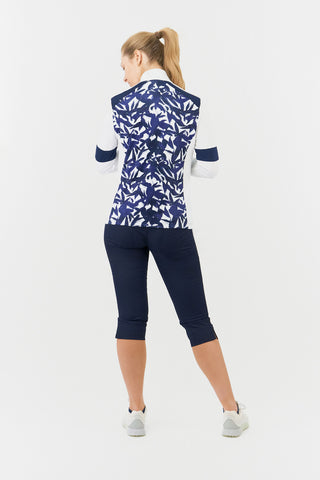 The Pure Golf Galena model shot showcasing the back of the mid-layer jacket in Wild Peony, featuring a botanical navy and white print on the body and inner neck, white sleeves, a navy band around the elbow, and a navy block across the shoulders. Designed for comfort and a flattering fit, it offers 4-way stretch to suit all body types. The jacket is anti-crease, anti-static, and quick-drying, and provides UPF 50+ sun protection. It is made from 95% Polyester and 5% Spandex. Paired with Navy Trust Capri's.