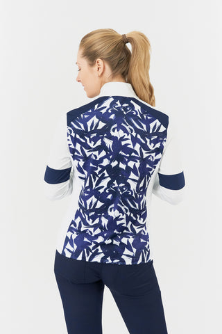 The Pure Golf Galena model shot features the back of the mid-layer jacket in Wild Peony, showcasing a botanical navy and white print on the body and inner neck, white sleeves, a navy band around the elbow, and a navy block across the shoulders. Designed for comfort and a flattering fit, the jacket offers 4-way stretch to suit all body types. It is anti-crease, anti-static, quick-drying, and provides UPF 50+ sun protection. Made from 95% Polyester and 5% Spandex.