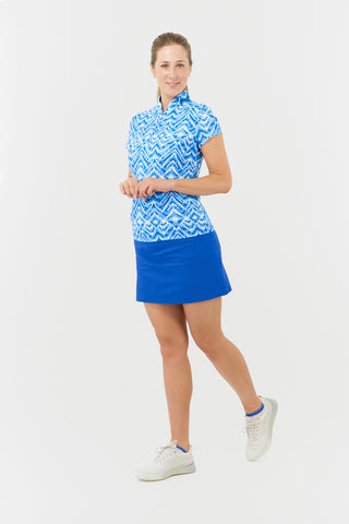 The front model shot of the Pure Golf Rise Santorini Skies Cap Sleeve Ladies' Golf Polo Shirt showcases a captivating Ikat-inspired design in shades of blue and white. With a regular fit and 4-way stretch, it offers a flattering shape for all body types. The mandarin collar adds an elegant touch. Made from 95% polyester and 5% spandex, it provides anti-crease, anti-static, and quick-dry properties, along with UPF 50+ sun protection. Paired with a Royal Blue Skort, it completes a stylish look. 