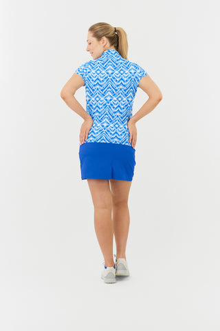 The back model shot of the Pure Golf Rise Santorini Skies Cap Sleeve Ladies' Golf Polo Shirt showcases a captivating Ikat-inspired design in shades of blue and white. With a regular fit and 4-way stretch, it offers a flattering shape for all body types. The mandarin collar adds an elegant touch. Made from 95% polyester and 5% spandex, it provides anti-crease, anti-static, and quick-dry properties, along with UPF 50+ sun protection. Paired with a Royal Blue Skort, it completes a stylish look. 