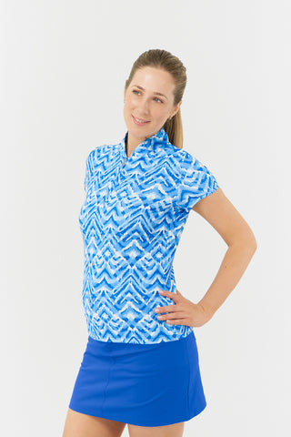 The front close-up model shot of the Pure Golf Rise Santorini Skies Cap Sleeve Ladies' Golf Polo Shirt showcases a captivating Ikat-inspired design in shades of blue and white. With a regular fit and 4-way stretch, it offers a flattering shape for all body types. The mandarin collar adds an elegant touch. Made from 95% polyester and 5% spandex, it provides anti-crease, anti-static, and quick-dry properties, along with UPF 50+ sun protection. Paired with a Royal Blue Skort, it completes a stylish look. 