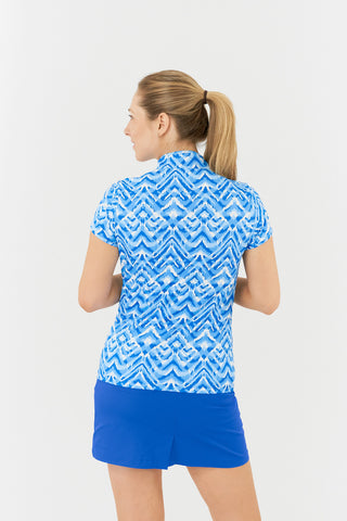 The Pure Golf Rise Santorini Skies cap sleeve Ladies' Golf Polo Shirt back model shot features a captivating Ikat-inspired design in shades of blue and white. With a regular fit and 4-way stretch, it provides both comfort and a flattering shape for all body types. The mandarin collar adds an elegant touch. Made from 95% polyester and 5% spandex, it offers anti-crease, anti-static, and quick-dry properties, along with UPF 50+ sun protection. Paired with a Royal Blue Skort.