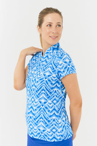 The close-up side model shot of the Pure Golf Rise Santorini Skies Cap Sleeve Ladies' Golf Polo Shirt showcases a captivating Ikat-inspired design in shades of blue and white. With a regular fit and 4-way stretch, it offers a flattering shape for all body types. The mandarin collar adds an elegant touch. Made from 95% polyester and 5% spandex, it provides anti-crease, anti-static, and quick-dry properties, along with UPF 50+ sun protection. Paired with a Royal Blue Skort to complete the look. 