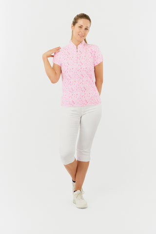 The Pure Golf Rise cap sleeve ladies' golf polo shirt in Soft Sakura, featured in a front model shot, showcases a subtle print of lilac, pink, and white petals against a light pink backdrop. Designed with a regular fit, it provides 4-way stretch for a flattering shape on all body types. The mandarin collar adds a touch of elegance. Made from 95% Polyester and 5% Spandex, the shirt offers anti-crease, anti-static, and quick-dry properties, along with UPF 50+ sun protection. Paired with White Trust Capri's. 