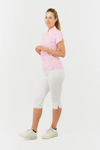 The Pure Golf Rise cap sleeve ladies' golf polo shirt in Soft Sakura, featured in a side model shot, showcases a subtle print of lilac, pink, and white petals against a light pink backdrop. Designed with a regular fit, it provides 4-way stretch for a flattering shape on all body types. The mandarin collar adds a touch of elegance. Made from 95% Polyester and 5% Spandex, the shirt offers anti-crease, anti-static, and quick-dry properties, along with UPF 50+ sun protection. White Trust Capri's. 