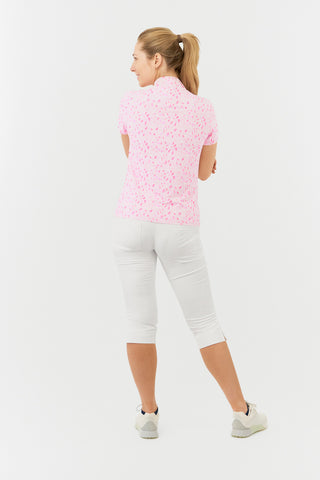 The Pure Golf Rise cap sleeve ladies' golf polo shirt in Soft Sakura, featured in a back model shot, showcases a subtle print of lilac, pink, and white petals against a light pink backdrop. Designed with a regular fit, it provides 4-way stretch for a flattering shape on all body types. The mandarin collar adds a touch of elegance. Made from 95% Polyester and 5% Spandex, the shirt offers anti-crease, anti-static, and quick-dry properties, along with UPF 50+ sun protection. White Trust Capri's. 