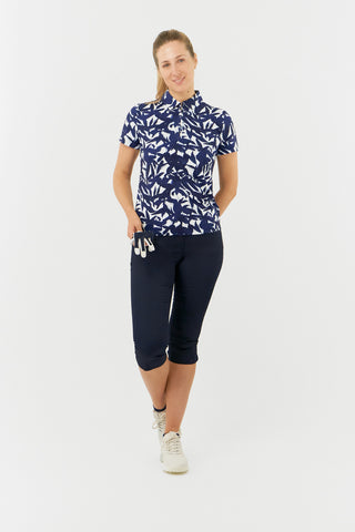 The Pure Golf Amity full length model shot showcases the short sleeve ladies' golf polo shirt, featuring a botanical print in Navy and white paired with Navy Trust Capri's. This regular-fit polo shirt offers a 4-way stretch for comfort and a flattering fit on all body types. With its traditional collar, it delivers a classic and stylish look. The shirt is designed to be anti-crease, anti-static, and quick-drying, with UPF 50+ protection. Made from 95% Polyester and 5% Spandex. 