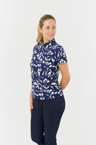 The Pure Golf Amity slight side model shot highlights the short-sleeve ladies' golf polo shirt, featuring a botanical print in navy and white, paired with navy golf trousers. This regular-fit polo shirt offers a 4-way stretch for comfort and a flattering fit on all body types. Its traditional collar adds a classic, stylish touch. The shirt is designed to be anti-crease, anti-static, and quick-drying, while also providing UPF 50+ protection. Made from 95% polyester and 5% spandex