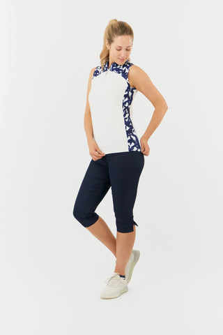 The Pure Golf Freda model side shot showcases the sleeveless ladies' golf polo shirt, featuring a botanical print in navy and white across the shoulders, sides, and mandarin collar, complemented by a white body. This regular-fit polo shirt offers a 4-way stretch for comfort and a flattering fit on all body types. Designed to be anti-crease, anti-static, and quick-drying, it also provides UPF 50+ protection. Made from 95% Polyester and 5% Spandex. Paired with Navy Trust Capri's. 