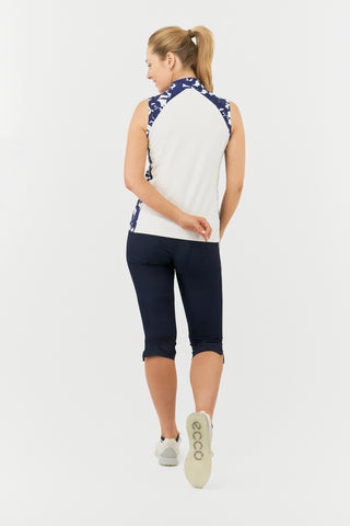 The Pure Golf Freda back view  lifestyle shot features the sleeveless ladies' golf polo shirt with a botanical print in navy and white across the shoulders, sides, and mandarin collar, with a white body. This regular-fit polo shirt provides a 4-way stretch for comfort and a flattering fit on all body types. It is designed to be anti-crease, anti-static, and quick-drying, while offering UPF 50+ protection. Made from 95% Polyester and 5% Spandex, it is paired with Navy Trust Capri's. 