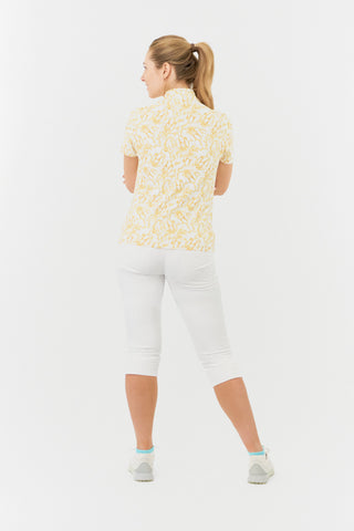 The Pure Golf Ellen short-sleeve ladies' polo shirt is a model shot back view, highlighting the sunkissed print and warm honey tones. It is paired with white Bermuda shorts. Designed for a regular fit, the polo provides a 4-way stretch for comfort and a flattering silhouette on all body types. The chic sweetheart collar adds a stylish flair. Made from 95% polyester and 5% spandex, the polo offers anti-crease, anti-static, and quick-dry benefits, along with UPF 50+ sun protection.