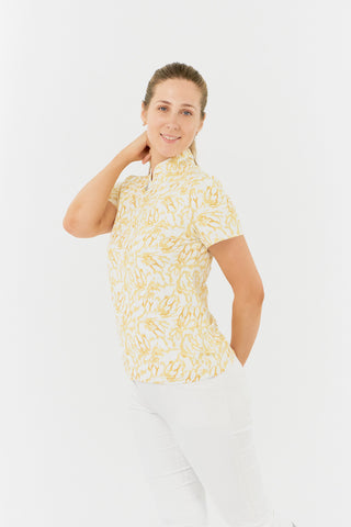 The Pure Golf Ellen short-sleeve ladies' polo shirt model shot is displayed in a subtle side view, showcasing the sunkissed print and soft honey tones. It is paired with white Bermuda shorts. Crafted with a regular fit, the polo offers a 4-way stretch for comfort and a flattering shape on all body types. The elegant sweetheart collar adds a touch of style. Made from 95% polyester and 5% spandex, the polo features anti-crease, anti-static, and quick-dry properties, as well as UPF 50+ sun protection.