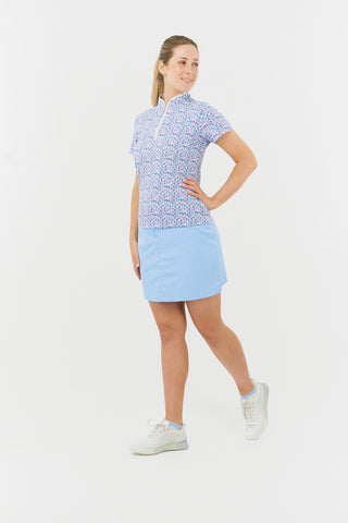 The Pure Golf Ellen short-sleeve ladies' polo shirt model shot full length side view showcasing our Over Eighteen Print featuring cocktail motifs in a subtle scale. Designed with a regular fit, it offers a 4-way stretch for comfort and a flattering shape on all body types. The stylish sweetheart collar adds a fashionable touch. This polo is made from 95% Polyester and 5% Spandex, offering anti-crease, anti-static, and quick-dry properties, along with UPF 50+ sun protection. Paired with a light blue skort. 