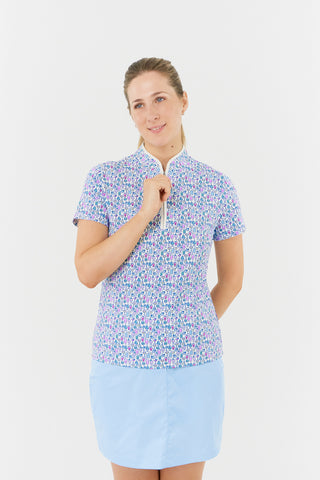 The Pure Golf Ellen short-sleeve ladies' polo shirt model shot front view showcasing our Over Eighteen Print featuring cocktail motifs in a subtle scale. Designed with a regular fit, it offers a 4-way stretch for comfort and a flattering shape on all body types. The stylish sweetheart collar adds a fashionable touch. This polo is made from 95% Polyester and 5% Spandex, offering anti-crease, anti-static, and quick-dry properties, along with UPF 50+ sun protection. Paired with a light blue skort. 