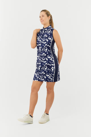 The Pure Golf model shot of the Heather Sleeveless dress features a botanical print in navy and white, accented by a flattering navy side panel. Made from 4-way stretch material for maximum flexibility and unrestricted movement, it also includes detachable shorts for added versatility and coverage. The mock neck collar offers a sleek, modern appearance while ensuring a flattering fit for all body types. Designed to be anti-crease, anti-static, and quick-drying, it also provides UPF 50+ protection.