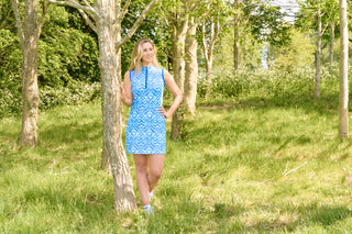 The Pure Golf lifestyle shot of the Sophie Sleeveless dress showcases the Santorini Skies print in shades of blue and white, accented by a flattering Royal Blue side panel. Made from 4-way stretch fabric, it ensures flexibility and freedom of movement, with detachable shorts for extra coverage. The mock neck collar offers a sleek, modern appearance, and the dress is designed to flatter all body types. Engineered to resist creasing, static, and moisture, it also provides UPF 50+ protection.