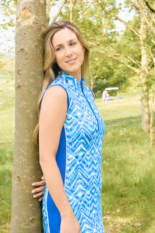 The Pure Golf side lifestyle shot of the Sophie Sleeveless dress showcases the Santorini Skies print in shades of blue and white, accented by a flattering Royal Blue side panel. Made from 4-way stretch fabric, it ensures flexibility and freedom of movement, with detachable shorts for extra coverage. The mock neck collar offers a sleek, modern appearance, and the dress is designed to flatter all body types. Engineered to resist creasing, static, and moisture, it also provides UPF 50+ protection.