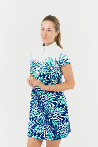 The Pure Golf Carly Cap Sleeve dress showcases an aquatic design in shades of blue and ocean hues, featuring a print of floating palms from the chest down in this close up model shot. This longer-length dress is crafted from 4-way stretch fabric for enhanced flexibility, with detachable shorts for added coverage. Its flattering fit suits all body types, while the mock neck collar delivers a sleek style. Designed to resist creasing, static, and moisture, the dress also offers UPF 50+ protection. 