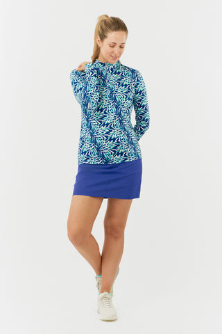 The Pure Golf model shot of the Serenity long-sleeve ladies' golf zip top showcases a soothing aquatic design in multi-blue and ocean tones, featuring a floating palms print. Designed with a regular fit, it offers 4-way stretch for comfort and a flattering shape on all body types. The mock neck collar adds a sleek, touch. Made from 95% Polyester and 5% Spandex, the top has anti-crease, anti-static, and quick-dry properties, along with UPF 50+ sun protection. Paired with a Royal Blue Skort.