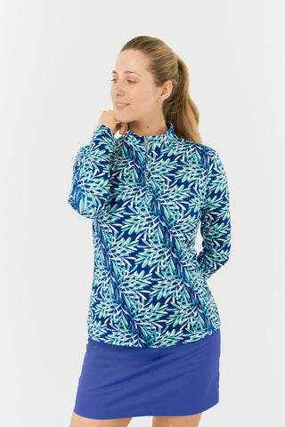 The Pure Golf model close up shot of the Serenity long-sleeve ladies' golf zip top showcases a soothing aquatic design in multi-blue and ocean tones, featuring a floating palms print. Designed with a regular fit, it offers 4-way stretch for comfort and a flattering shape on all body types. The mock neck collar adds a sleek, touch. Made from 95% Polyester and 5% Spandex, the top has anti-crease, anti-static, and quick-dry properties, along with UPF 50+ sun protection. Paired with a Royal Blue Skort.