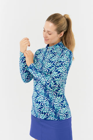 The Pure Golf side model shot of the Serenity long-sleeve ladies' golf zip top showcases a soothing aquatic design in multi-blue and ocean tones, featuring a floating palms print. Designed with a regular fit, it offers 4-way stretch for comfort and a flattering shape on all body types. The mock neck collar adds a sleek, touch. Made from 95% Polyester and 5% Spandex, the top has anti-crease, anti-static, and quick-dry properties, along with UPF 50+ sun protection. Paired with a Royal Blue Skort.