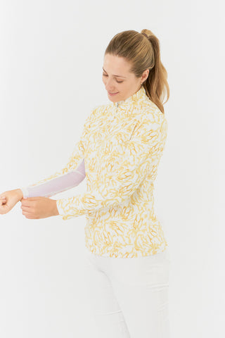 A side model shot of the Pure Golf Balance Sun Kissed Long Sleeve Ladies' Golf Zip Top from our Ray Flower collection features a beautiful print in warm honey hues. With a regular fit and 4-way stretch, it ensures comfort for all body types. The mock neck collar adds a sleek look, while mesh sleeve panels in white enhance breathability. Crafted from 95% polyester and 5% spandex, it offers anti-crease, anti-static, and quick-dry properties, along with UPF 50+ protection. Paired with White Trust Capri's.